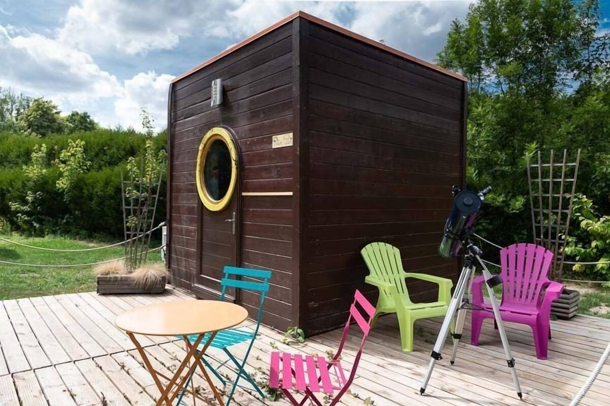 Eine Holzhütte mit rundem Fenster, einem Teleskop und bunten Adirondack-Stühlen auf einer Holzterrasse.