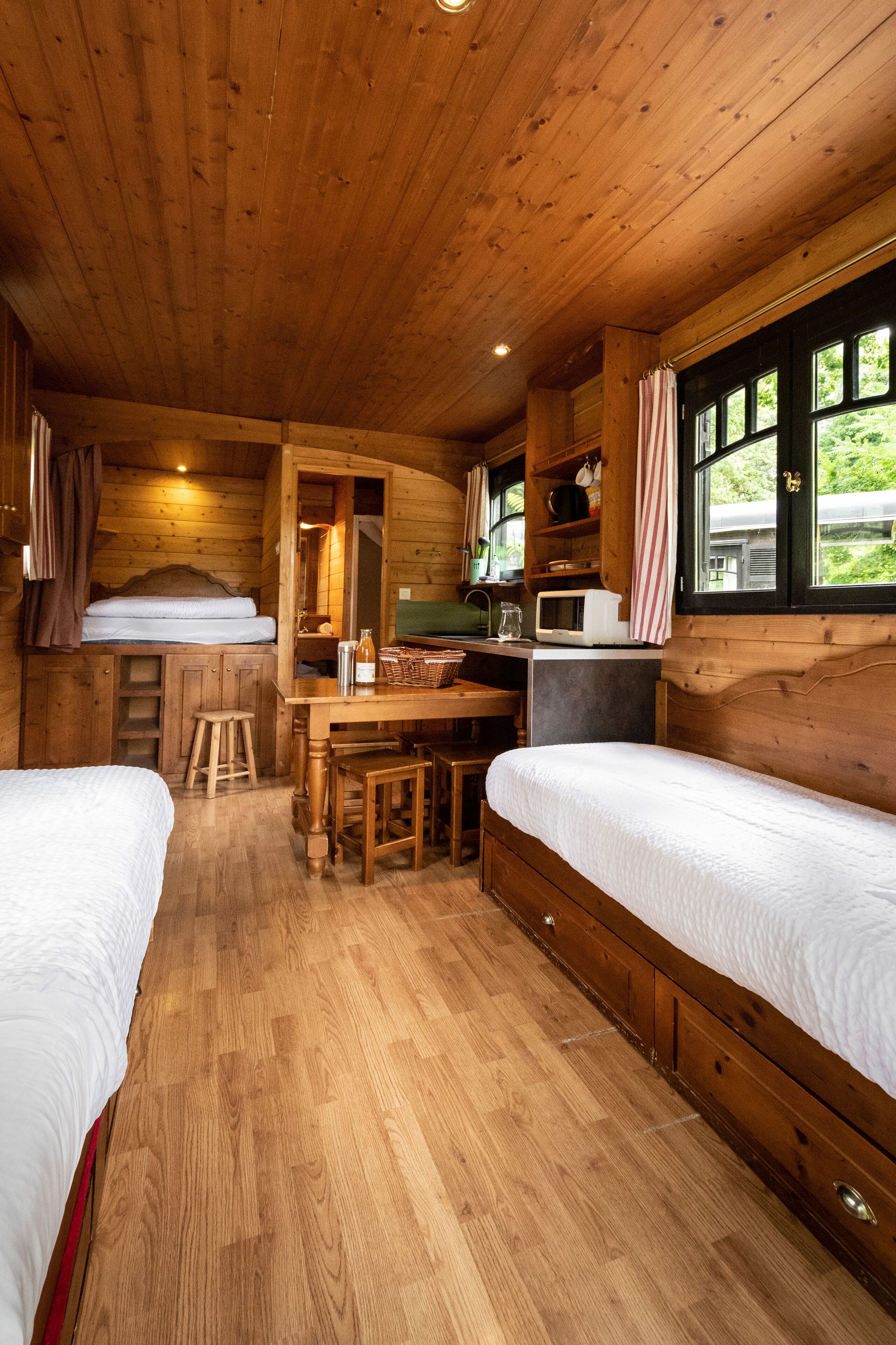 Une cabane comprenant deux lits, une table à manger en bois, un coin cuisine et une fenêtre avec des rideaux.