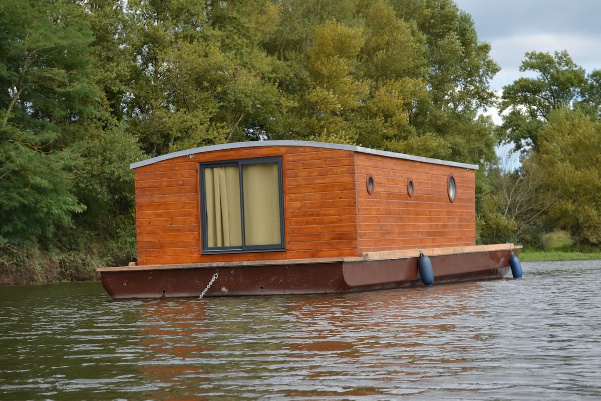 Ein schwimmendes Hausboot aus Holz mit Fenster und Tür, umgeben von Wasser und Bäumen.