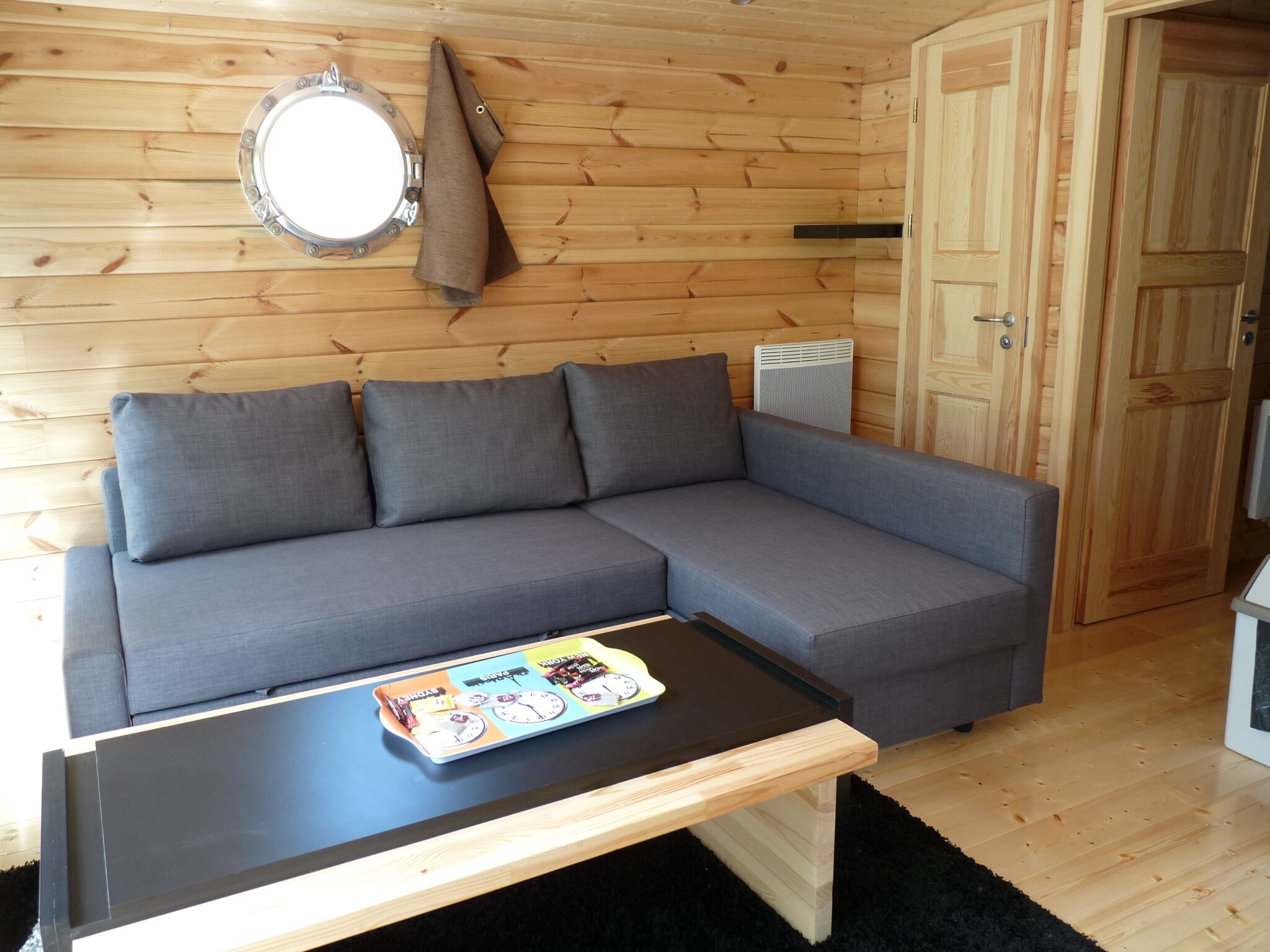 Interior moderno de una cabaña de troncos con un sofá seccional gris, una mesa de madera con un juego de mesa, un espejo redondo y una puerta.