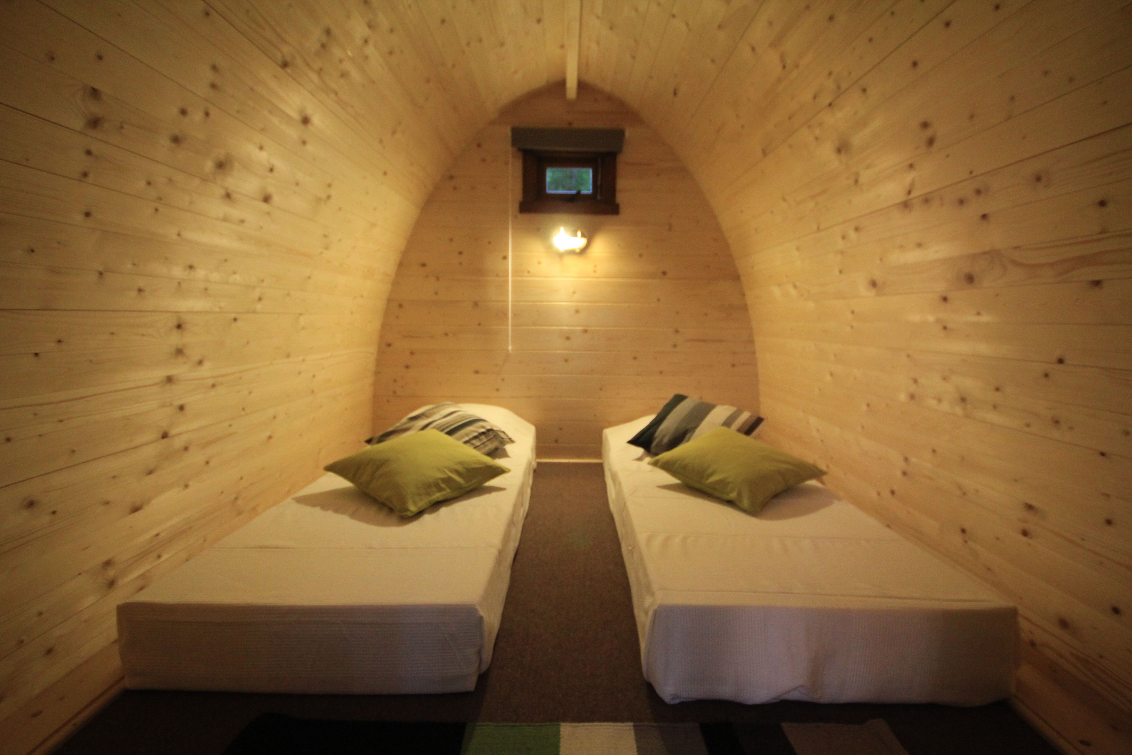 A small, cozy room with two beds, each with pillows and a small table. A light is visible on the wall above the beds.