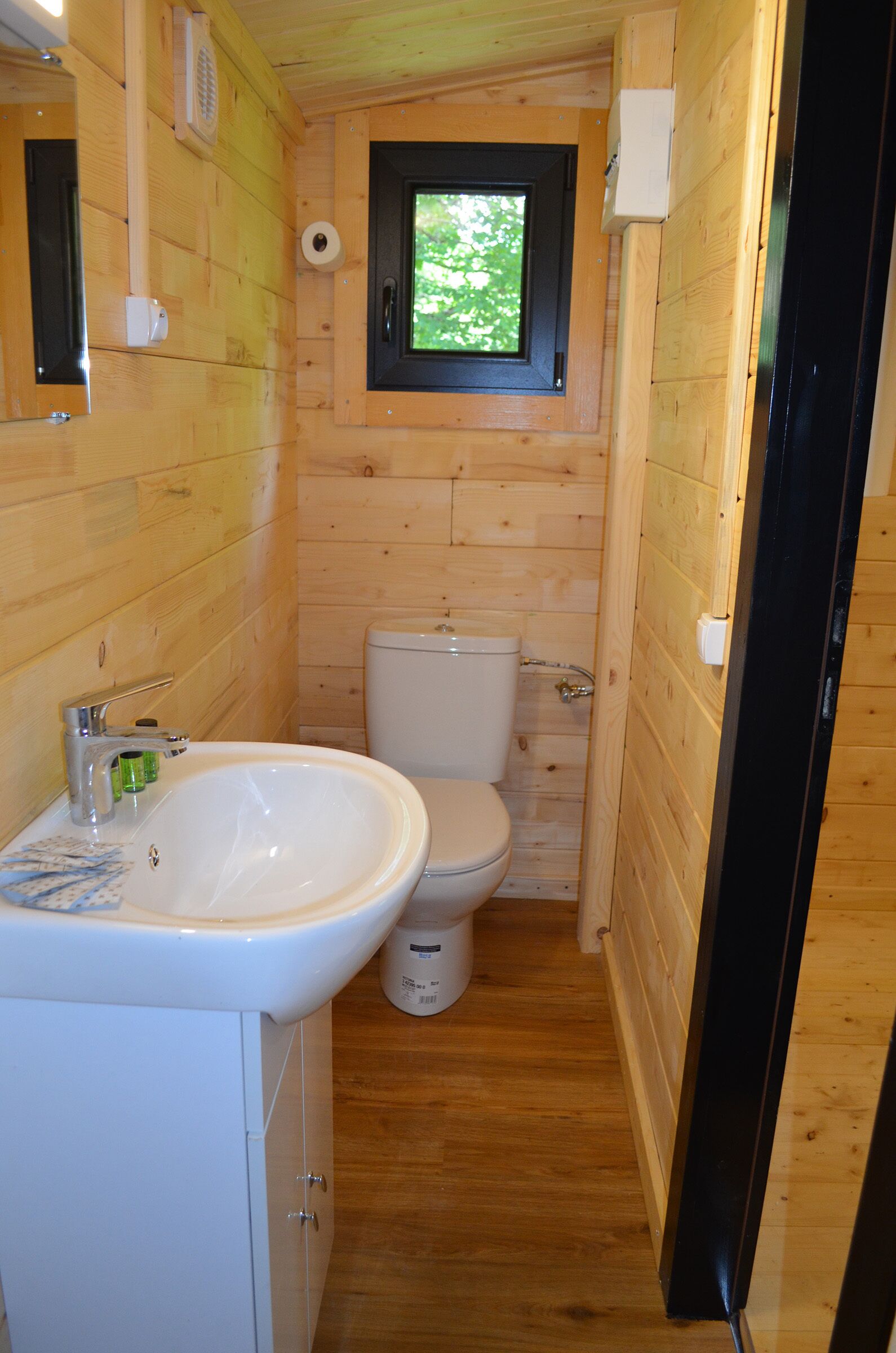 A compact wooden bathroom with a white sink, toilet, and a small window above the toilet.