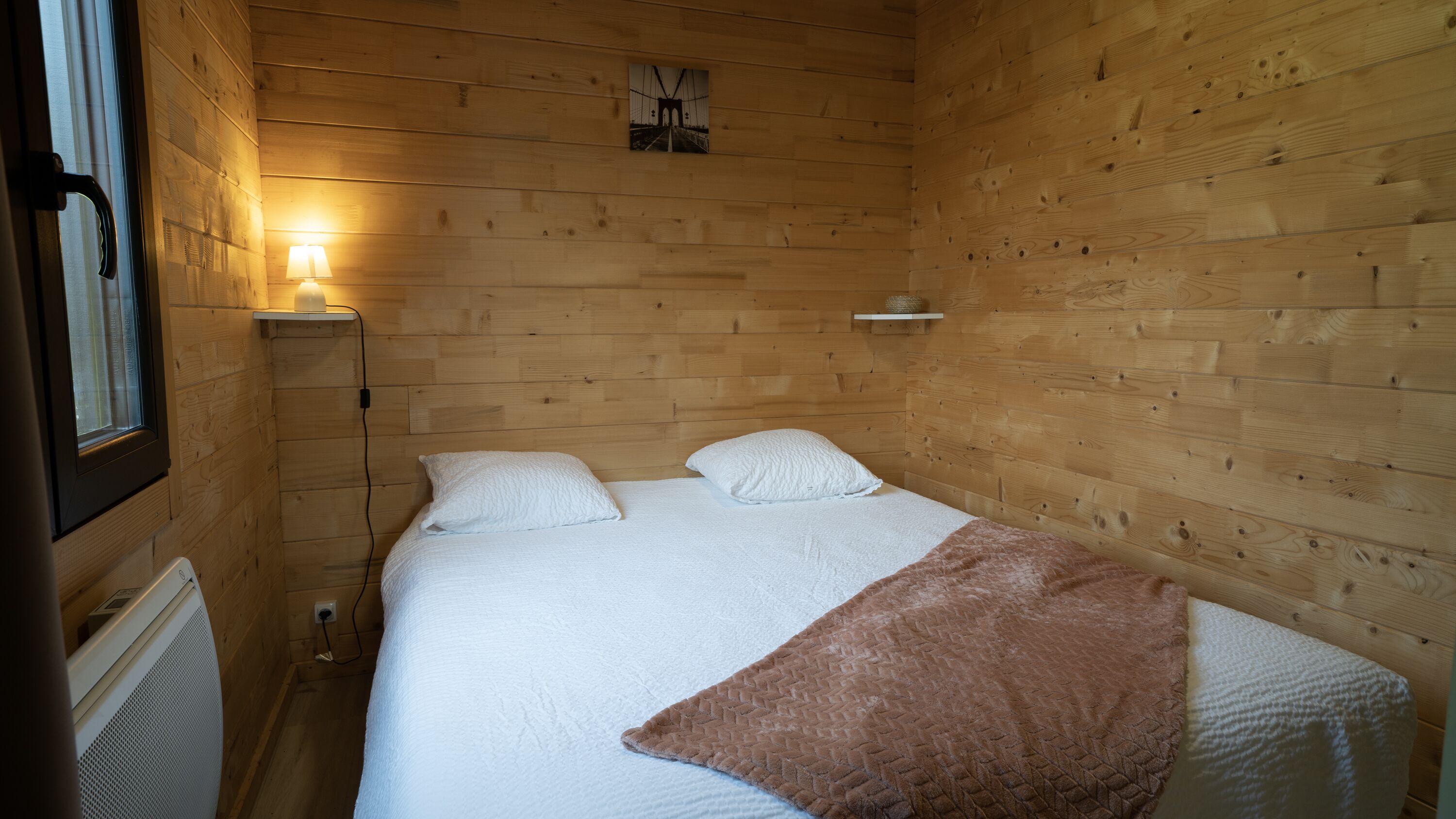Une pièce en bois avec un lit, une table de chevet, une lampe et une petite étagère.