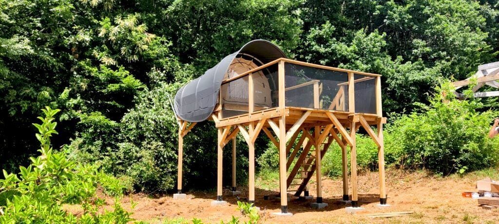 A wooden structure with a covered area, possibly a gazebo or small pavilion, surrounded by dense greenery.