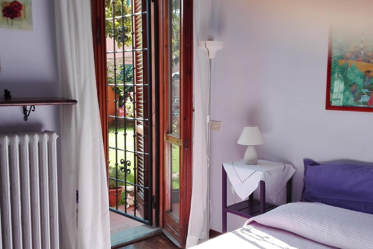 A bedroom with a bed, a nightstand with a lamp, a radiator, and a window with a view of the garden.