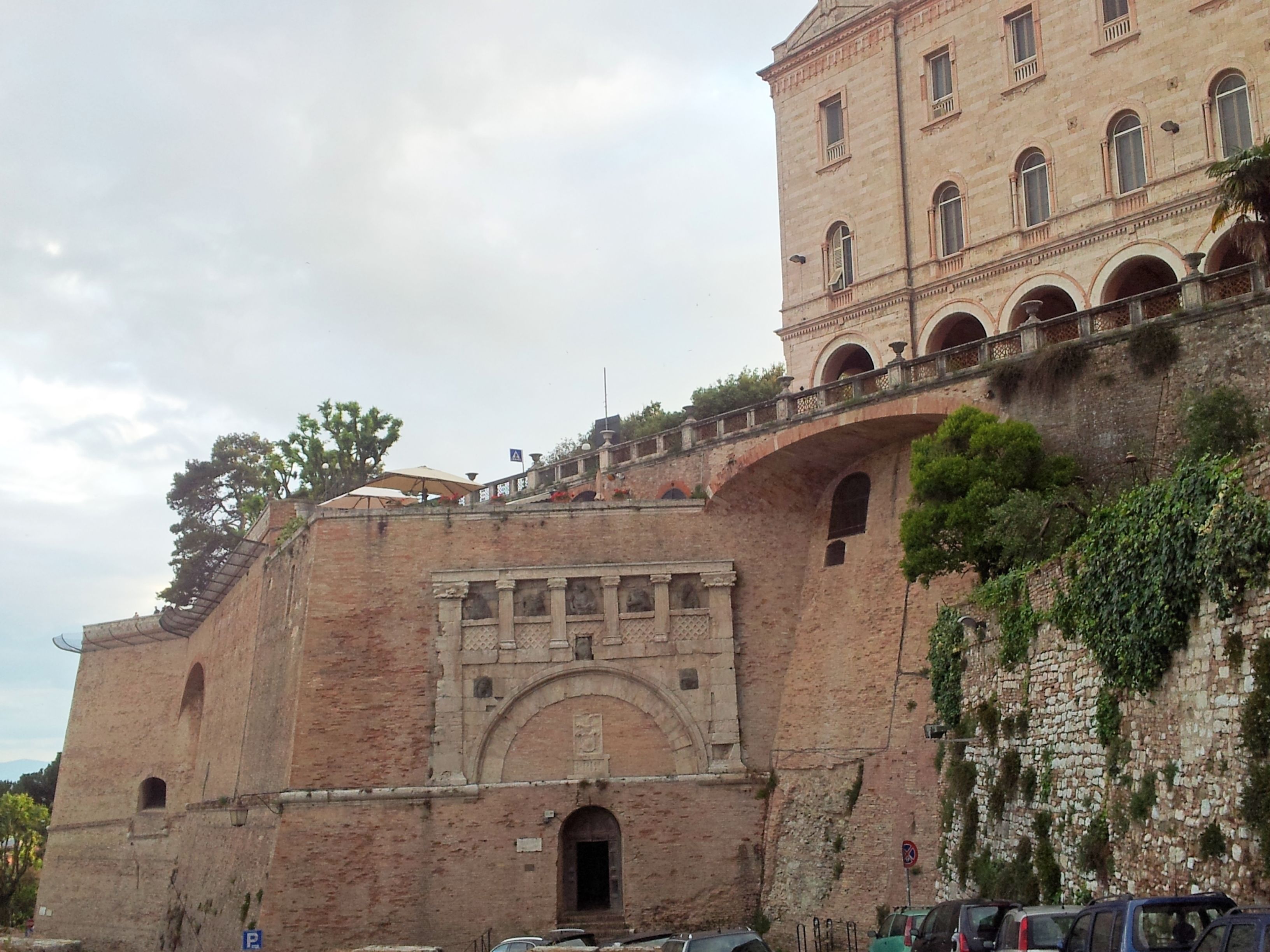 Un edificio storico con un'ampia entrata ad arco, un balcone con ringhiera e un muro in mattoni ricoperto di vegetazione.