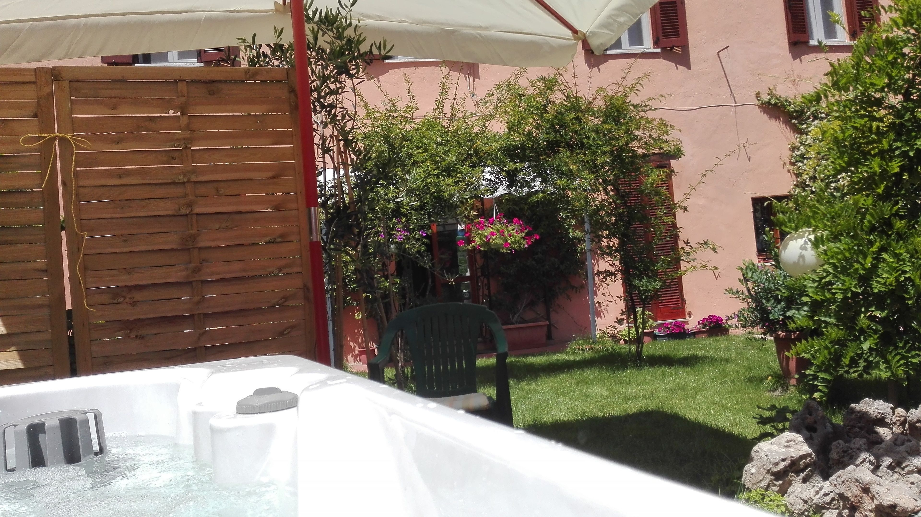 Area con vasca idromassaggio, recinzione in legno, una sedia verde e un giardino con piante e fiori in vaso.