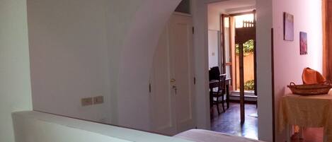 A room with a white bed, a table with a basket, and a door leading to a dining area with a chair and table.