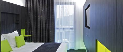 A modern hotel room with a large bed, green and white pillows, a small table, and a large window with sheer curtains.