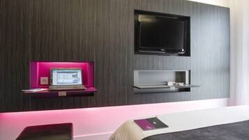 A modern hotel room with a flat-screen TV mounted on the wall, a laptop on a pink shelf, and a bedside table with a book.