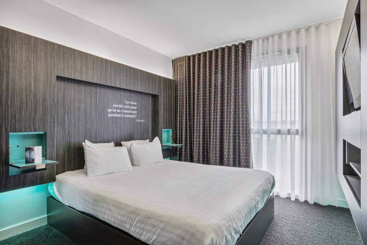 A modern hotel room with a large bed, a wall-mounted quote, a bedside table with a lamp, and a window with curtains.