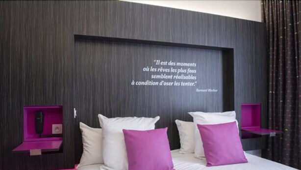 Quarto moderno com cama grande, almofadas brancas e roxas, e uma frase na parede.