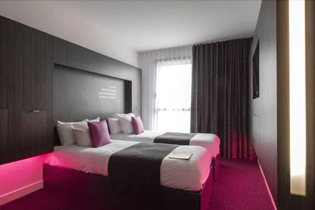 A modern hotel room with two beds, a large window, and a dark wood panel wall.