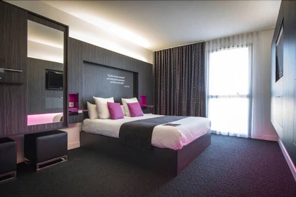 A modern hotel room with a large bed, a flat-screen TV mounted on the wall, a bedside table, and a large window with curtains.