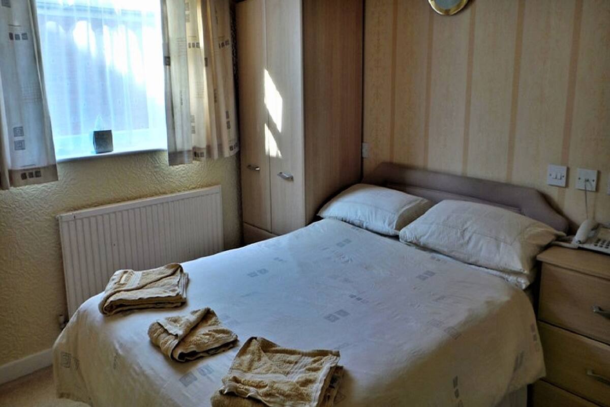 Ein Hotelzimmer mit Bett, Nachttisch, Heizkörper, Fenster mit Vorhängen und einer Wanduhr.