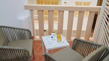 A balcony with two wicker chairs, a small table with a bottle and glass, and a view of a building with yellow shutters.