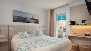 Habitación de hotel con cama, un cuadro de paisaje costero, escritorio con cafetera y taburetes de mimbre.