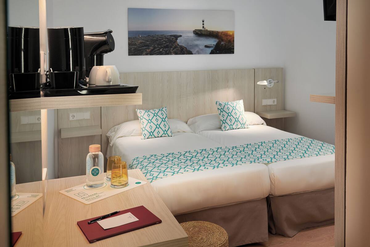 Una habitación de hotel con cama, mesita de noche y escritorio con cafetera y una botella.