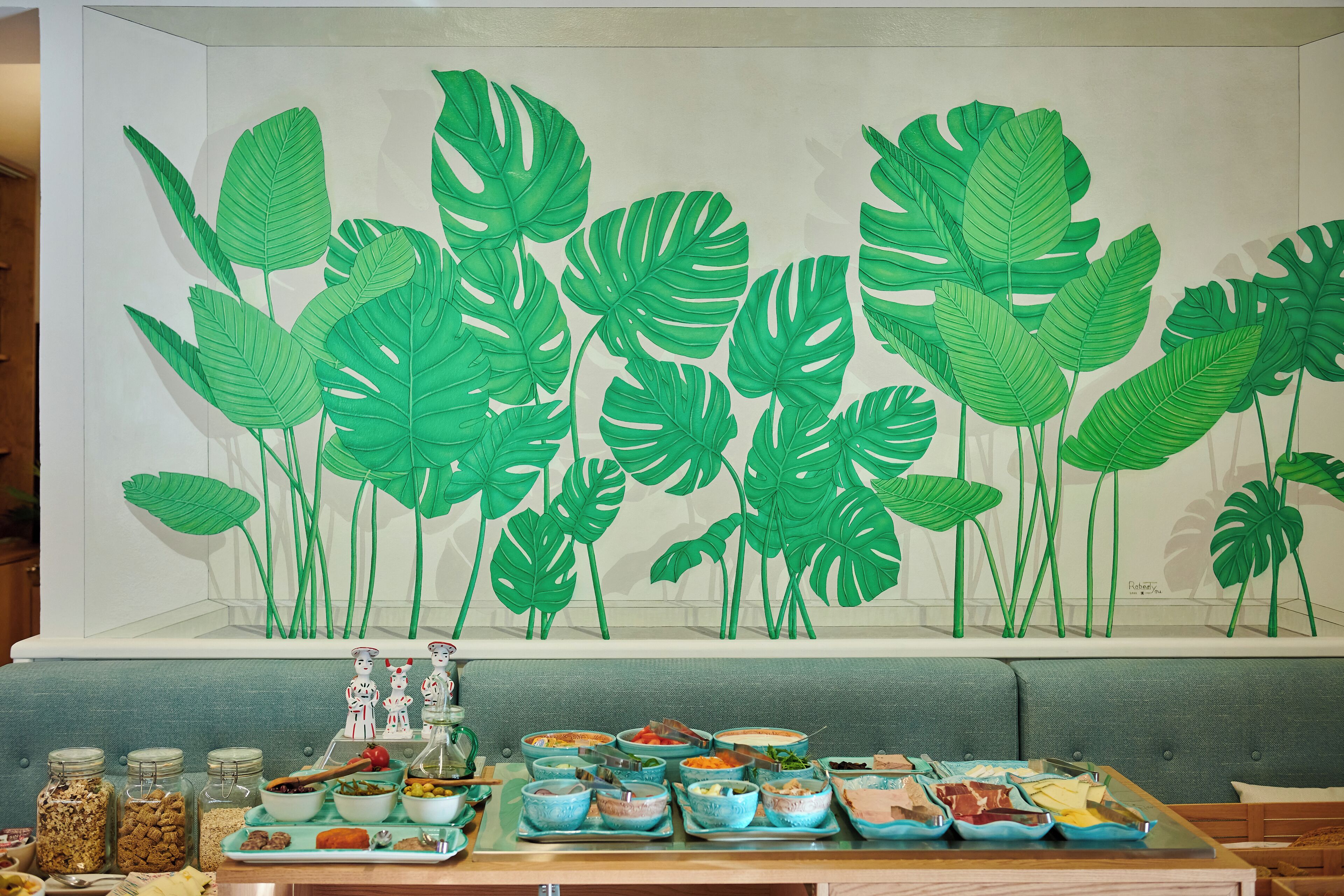A dining area with a wall mural of green leaves, a buffet table with various dishes, and a seating area with a grey sofa.