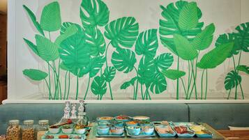 A dining area with a wall mural of green leaves, a buffet table with various dishes, and a seating area with a grey sofa.