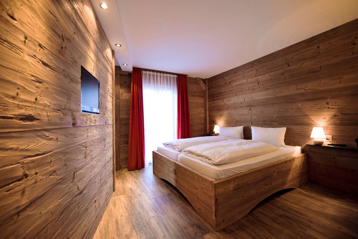 A wooden-paneled bedroom with a bed, two bedside lamps, and a window with red curtains.