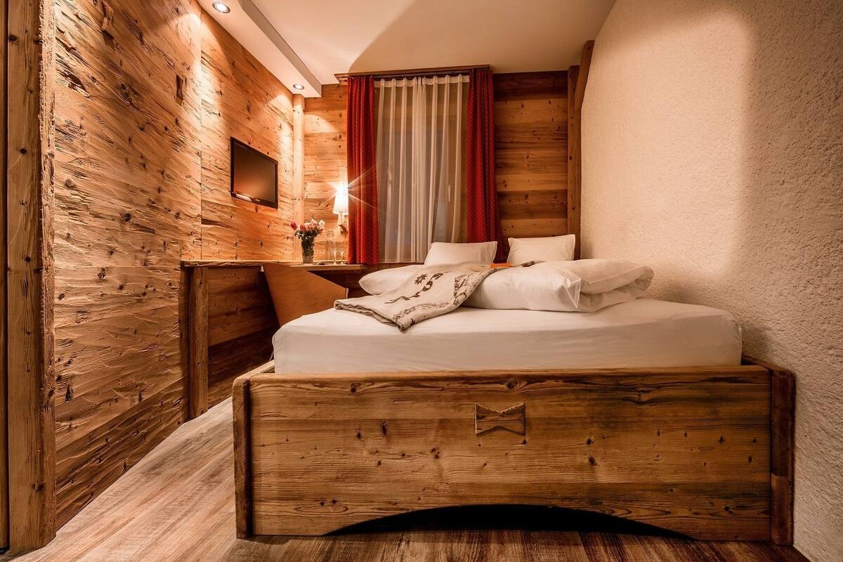 A wooden cabin-style bedroom with a bed, a nightstand, a TV, and a window with curtains.