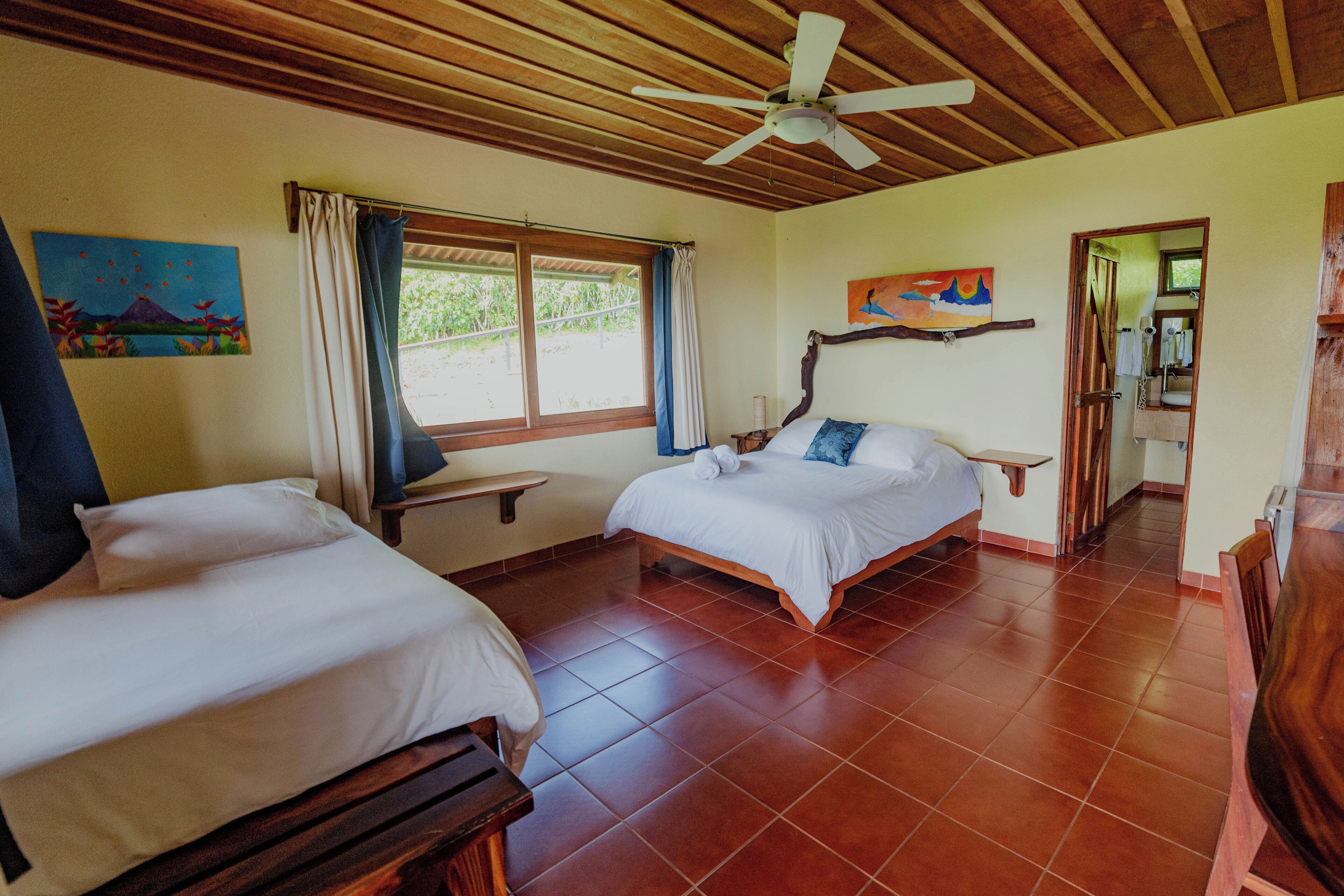 A room with two beds, a ceiling fan, a window with curtains, and artwork on the walls.