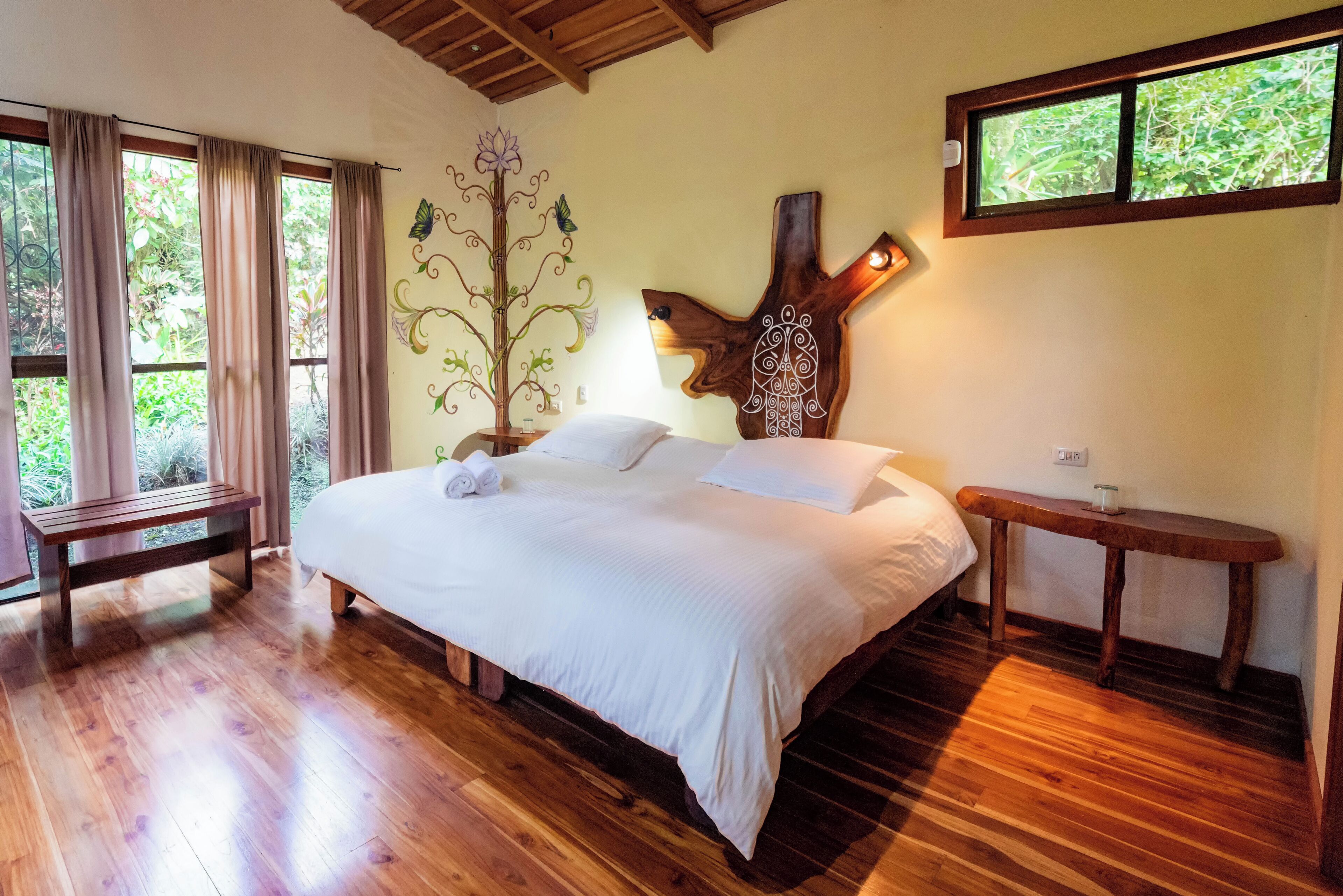 Un dormitorio con una cama grande, piso de madera, un banco, un árbol decorativo y una ventana con vistas al verdor.