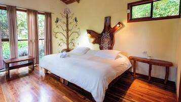 Un dormitorio con una cama grande, piso de madera, un banco, un árbol decorativo y una ventana con vistas al verdor.