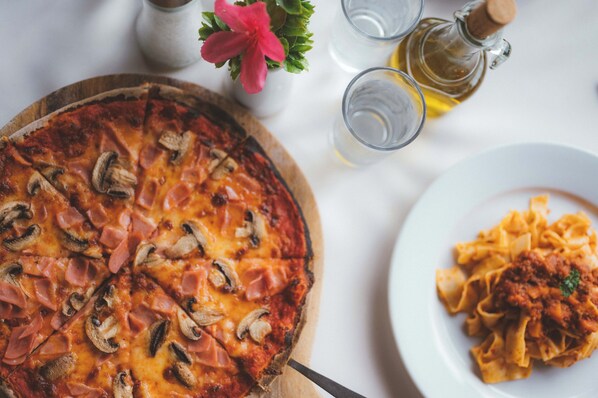 A pizza with ham and mushrooms on a wooden board, a plate of pasta with tomato sauce, and a small flower in a white vase. - Hotel Mystica (Santa Rosa)