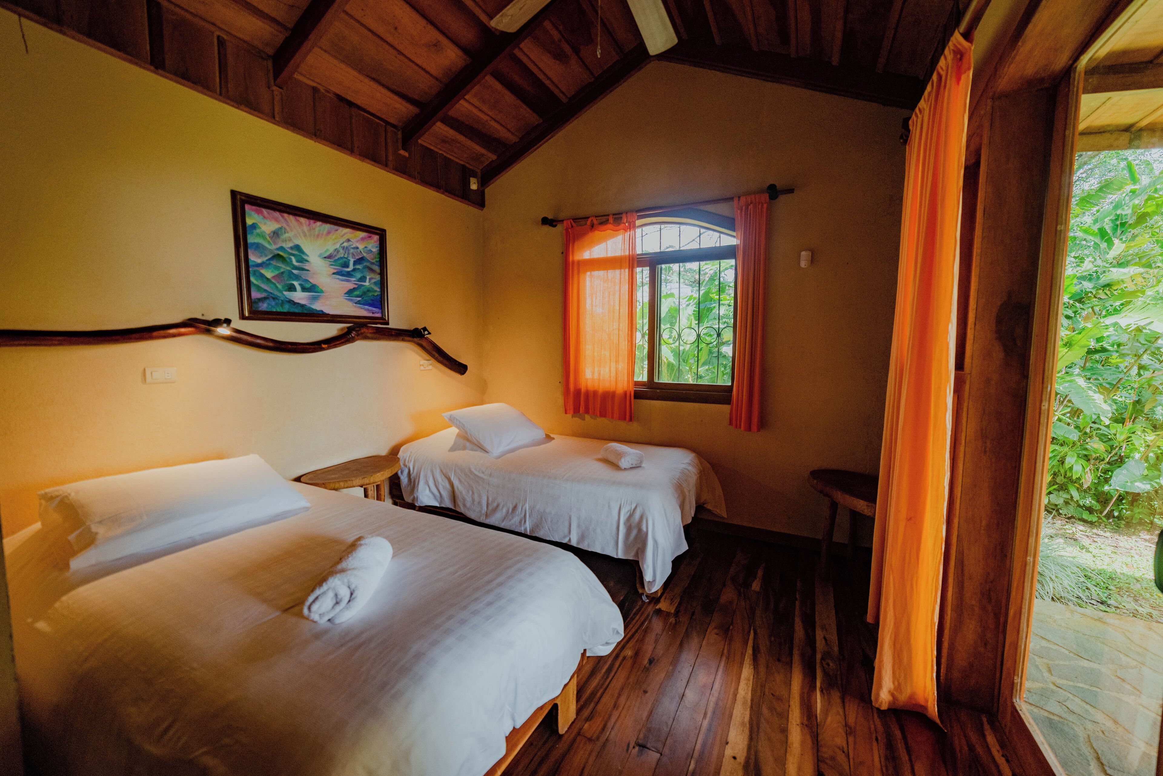 A room with two beds, wooden flooring, a painting on the wall, and a window with curtains.