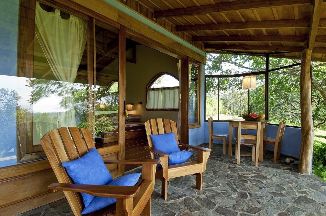 A wooden deck with two wooden chairs and a table, stone flooring, and large windows with curtains.