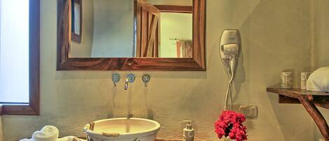 A bathroom with a wooden-framed mirror, a white sink with a floral design, a towel, a hairdryer, and a vase with pink flowers.