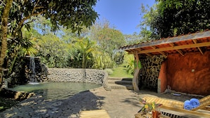 A stone patio with a small waterfall, a wooden lounge chair, a table with a drink and flowers, and a covered area with a red roof. - Hotel Mystica (Santa Rosa)