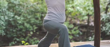 A person performing a yoga pose on a mat under a wooden structure with a natural backdrop.