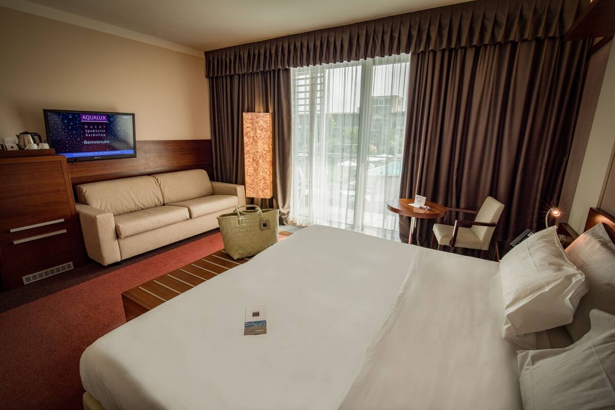 Quarto de hotel com cama grande, sofá, televisão, luminária e vista para o exterior.