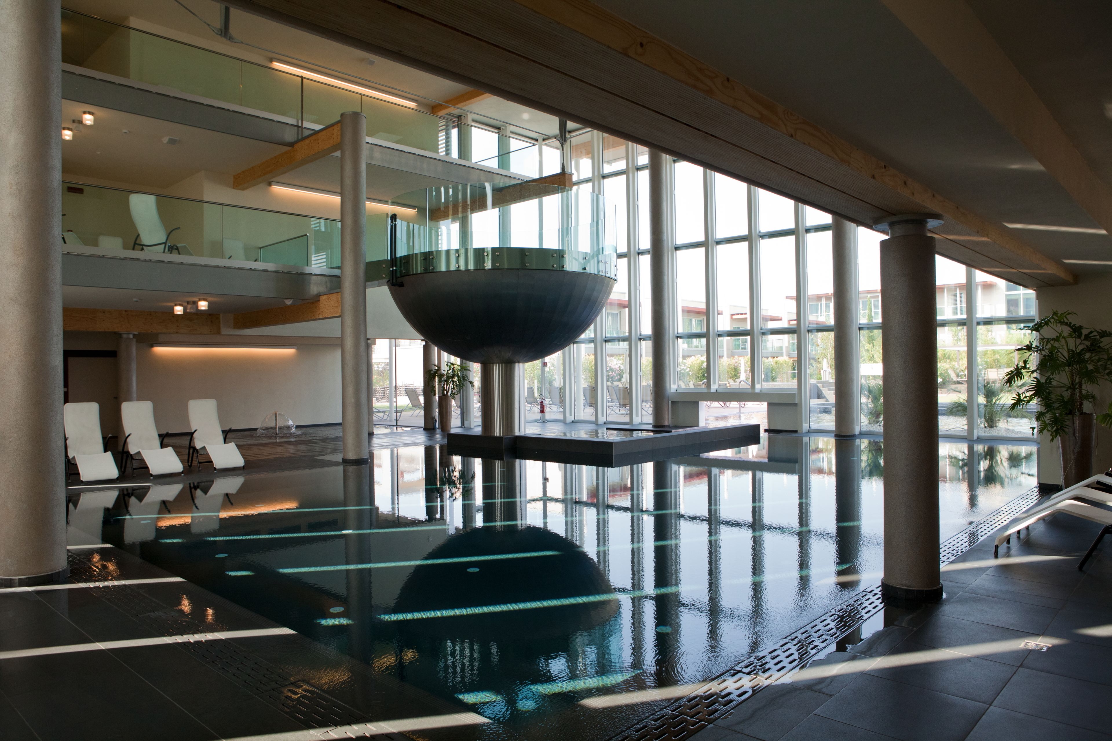 A modern indoor pool area with glass walls, a central bowl, and seating on the sides.
