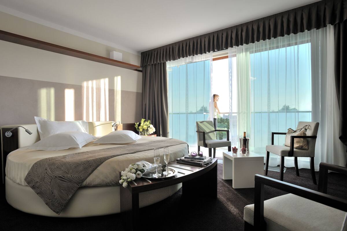 Quarto de hotel com uma cama grande, mesa de cabeceira com um serviço de objetos, uma cadeira, uma pequena mesa e varanda com vista.