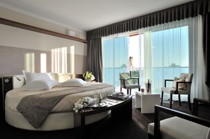 Habitación de hotel con una cama grande, una mesita de noche con una bandeja de objetos, una silla, una pequeña mesa y un balcón con vistas.