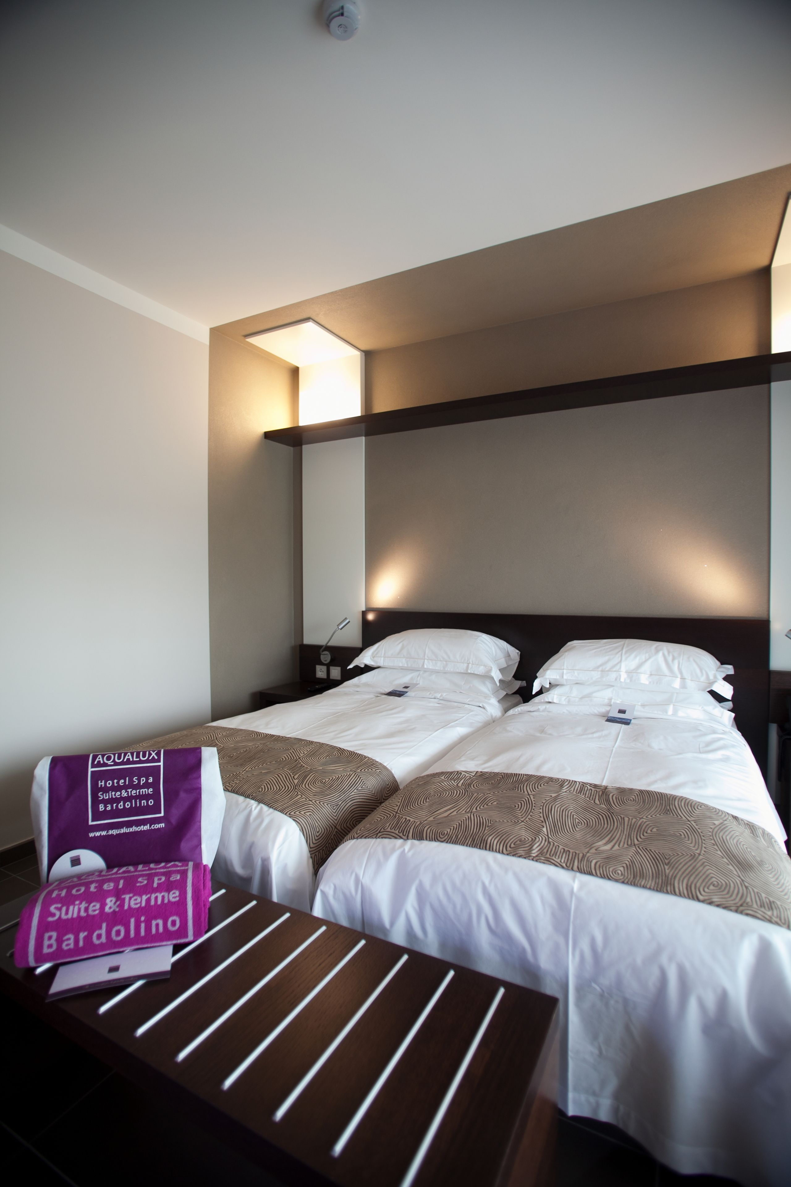 Una camera d'albergo con due letti, un comodino e una lampada fissata al muro.