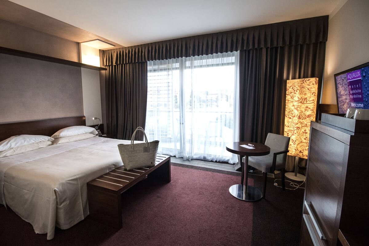 Quarto de hotel com cama, escrivaninha, cadeira, televisão e janela com cortinas.