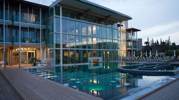 Un hotel moderno con fachada de vidrio, zona de piscina y mobiliario exterior.