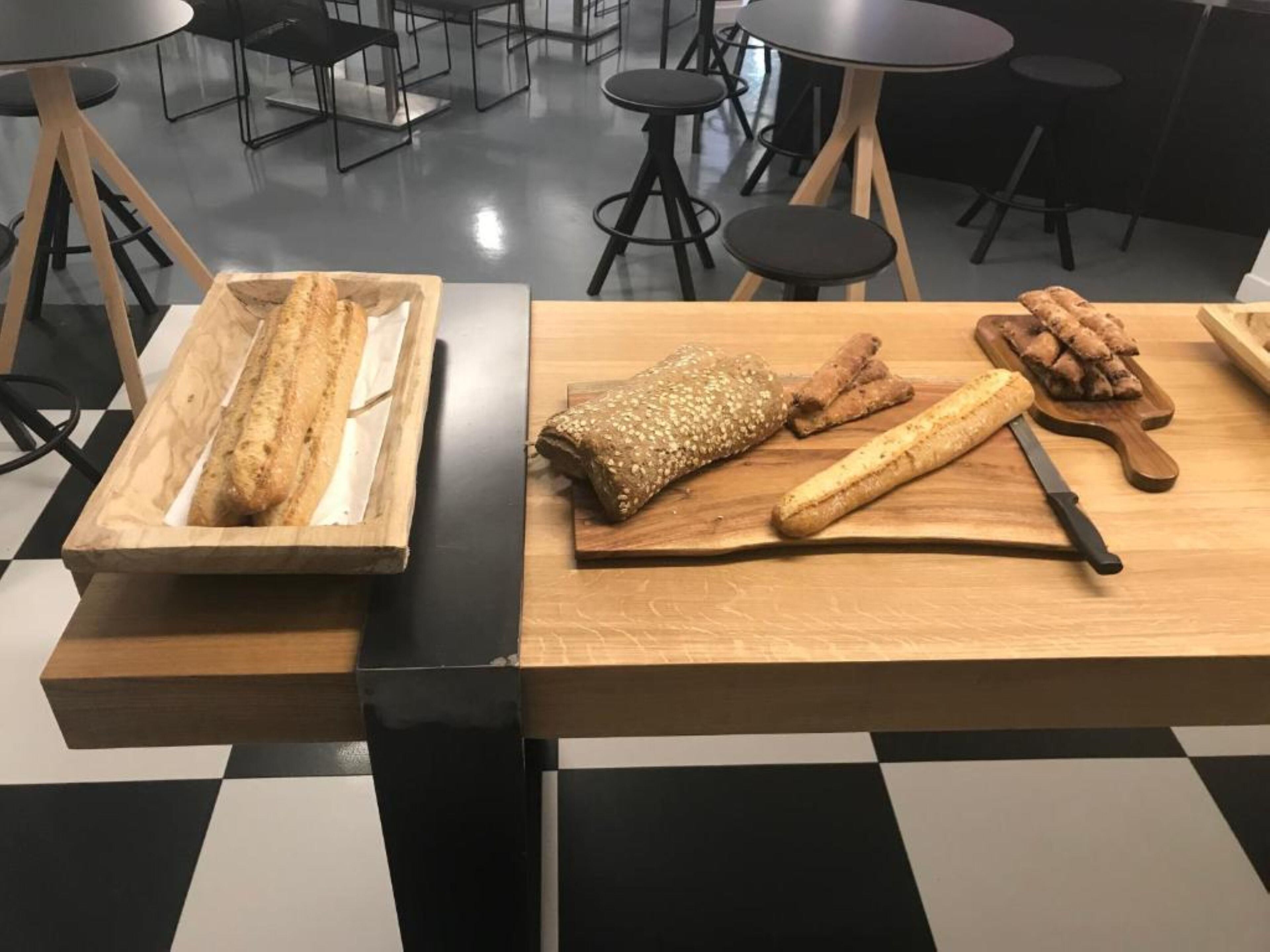 Un comedor con piso a cuadros, una mesa de madera con pan y un cuchillo, y sillas negras.
