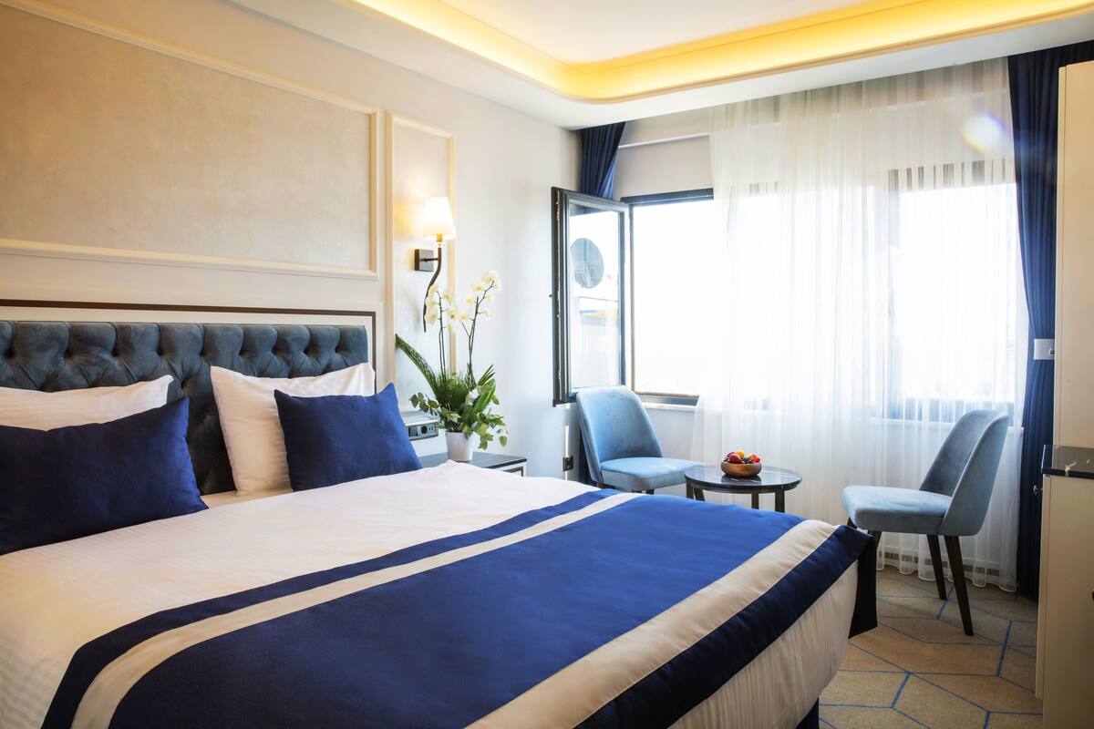 Una habitació d'hotel amb un llit gran, dues cadires, una taula petita amb fruita i una finestra amb cortines.
