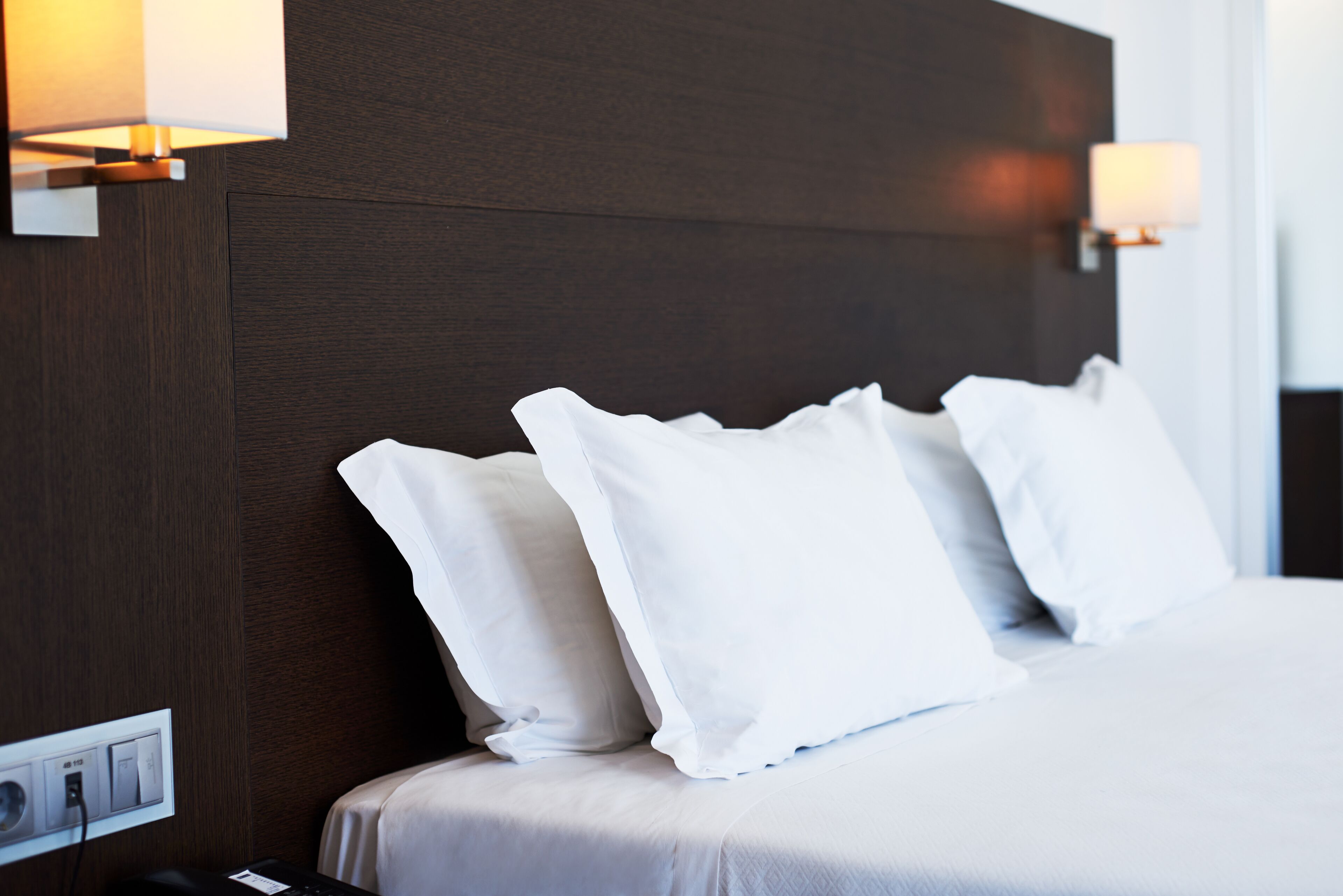 Una habitación de hotel con cabecera de madera oscura, ropa de cama blanca y lámparas fijadas a la pared.