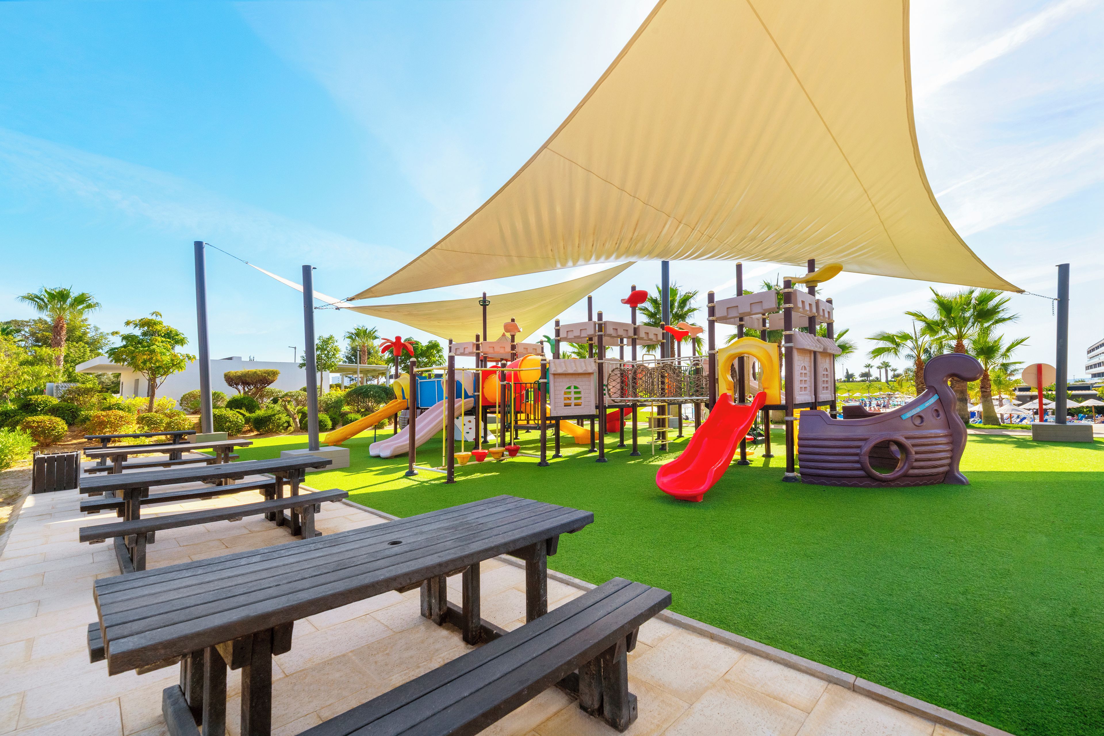 A playground with slides and climbing structures, surrounded by picnic tables and artificial grass.