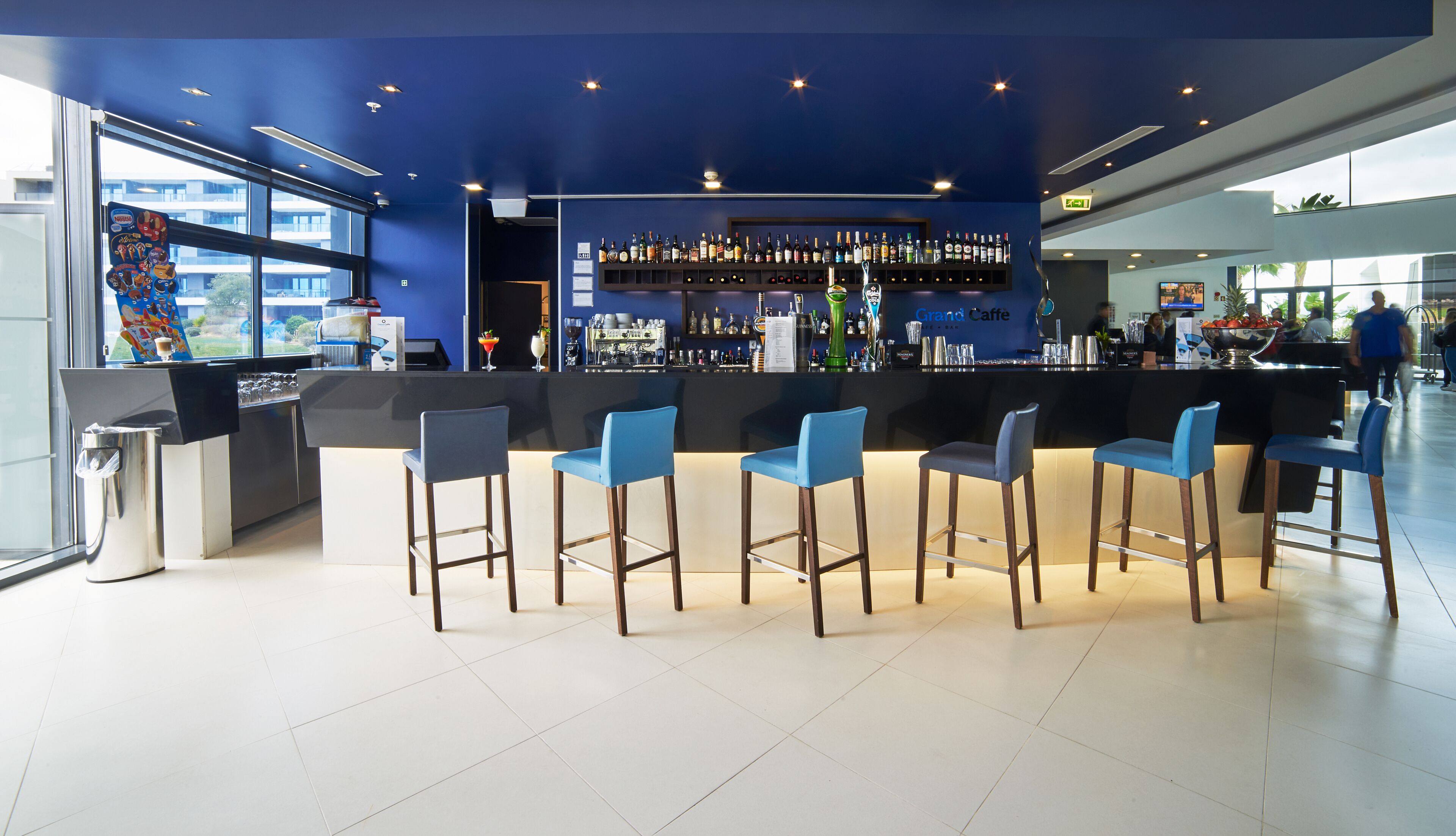 A modern bar area with a blue theme, featuring a counter with a variety of liquor bottles and a selection of glasses. There are five blue bar stools arranged around the counter.