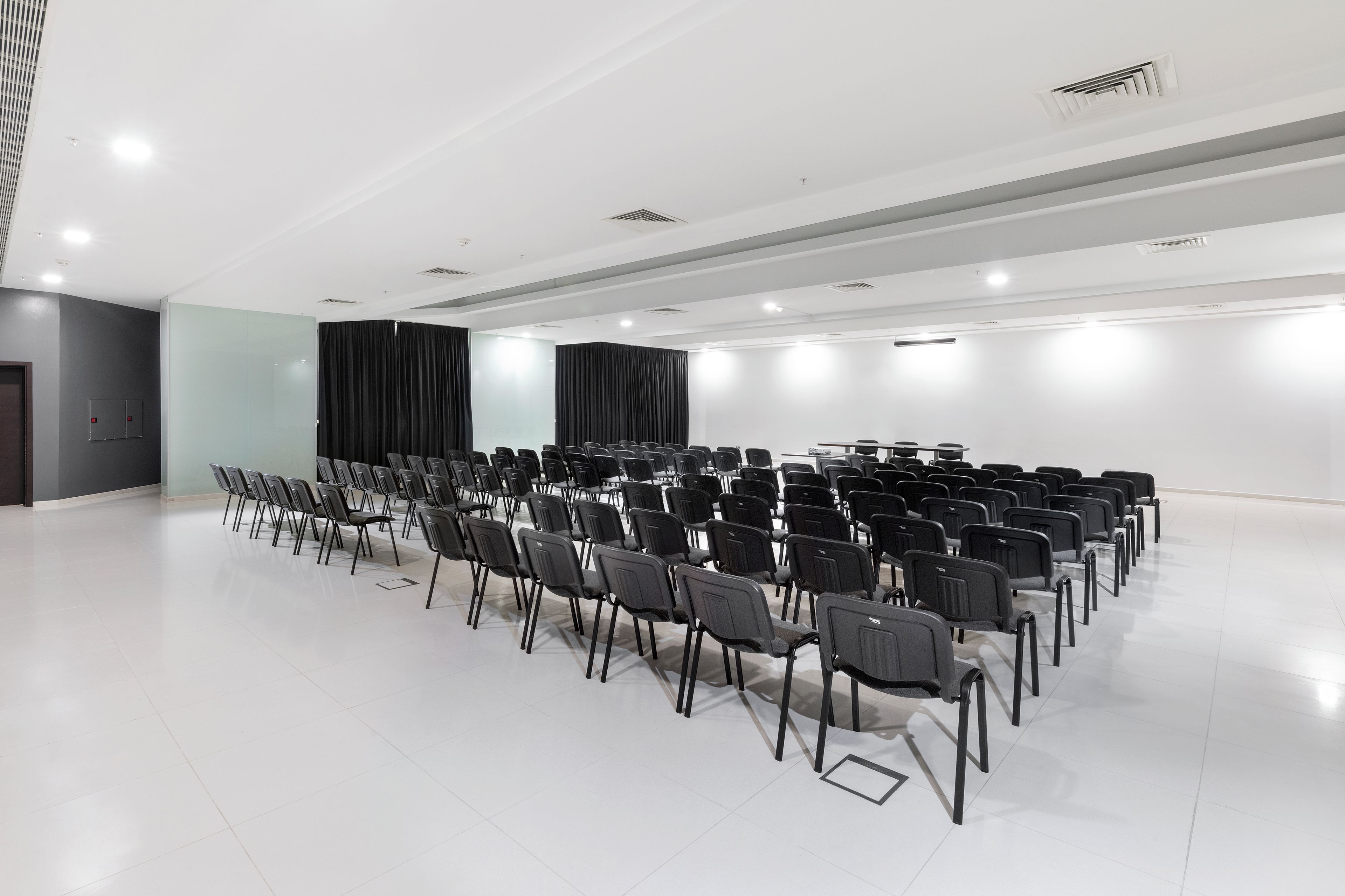 A conference room with rows of chairs, a stage with curtains, and a white ceiling with recessed lighting.