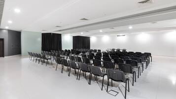 A conference room with rows of chairs, a stage with curtains, and a white ceiling with recessed lighting.