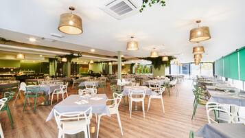 A spacious restaurant with multiple dining tables and chairs, a wooden floor, and hanging wicker lamps.